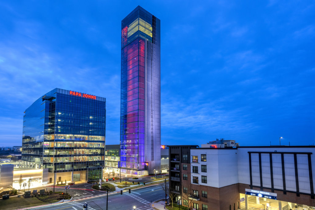 the TK elevator test tower is lit up at dusk. An office building sits to its left housing the Papa John's headquarters. 