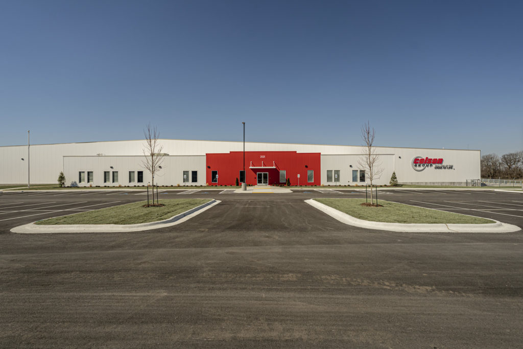 An exterior of a manufacturing facility. About three quarters of the facility's exterior walls are ivory, and a center portion is bright read. The Colson Group logo, which is text, is visible on the right side of the building. The building faces a parking lot.