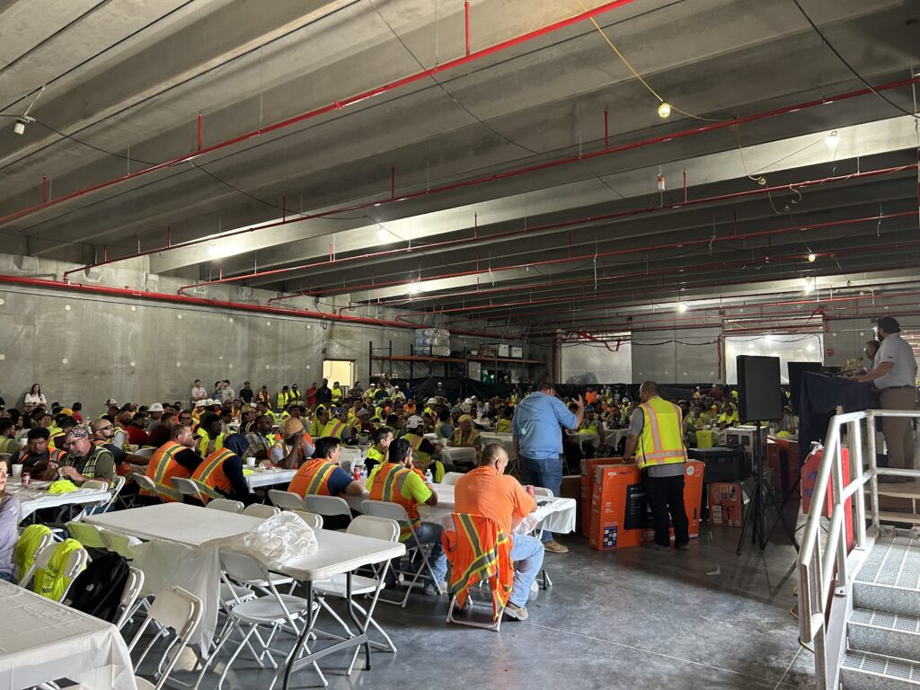 Dozens of construction workers gather for a lunch celebration inside of a facility. They wear high-visibility orange and yellow clothing.