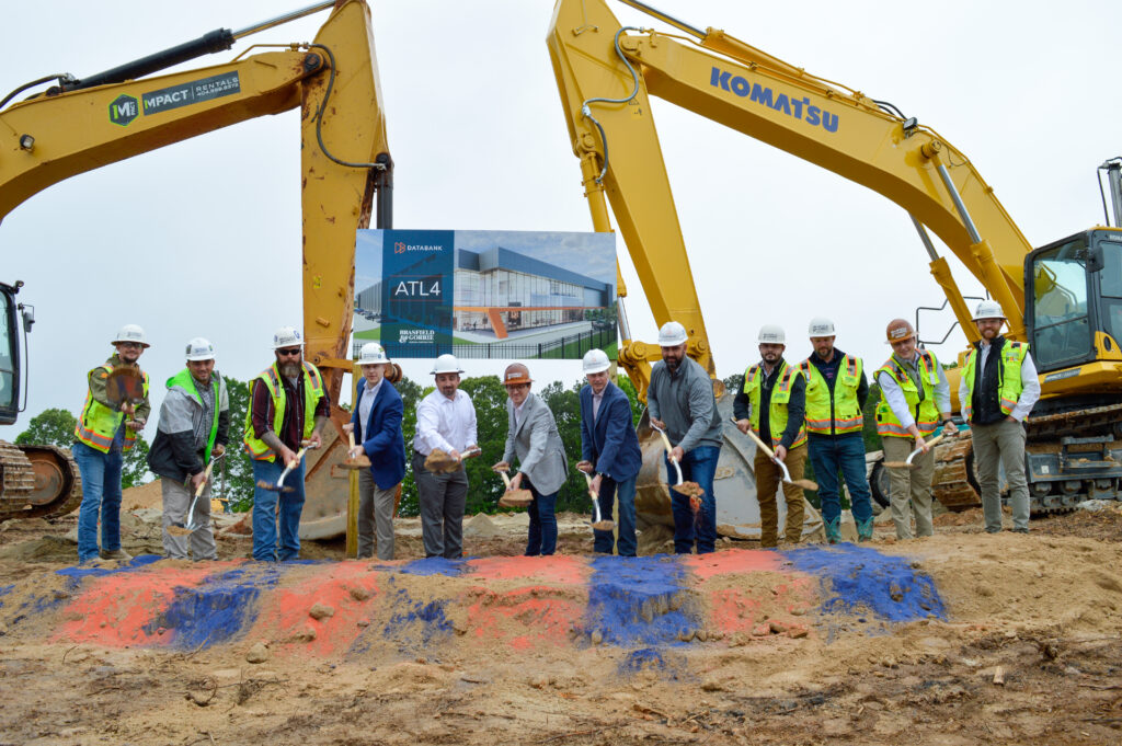 A group of contruction workers break ground in front of two pieces of yellow heavy machinery