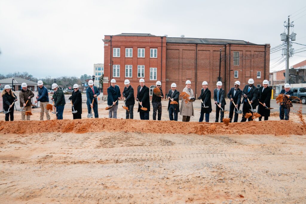 A line of 18 people in hardhats and holding shovels toss red dirt during a ceremonial groundbreaking
