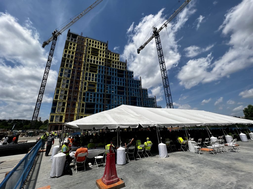 People gather under a white event tent in front of a residential tower under construction