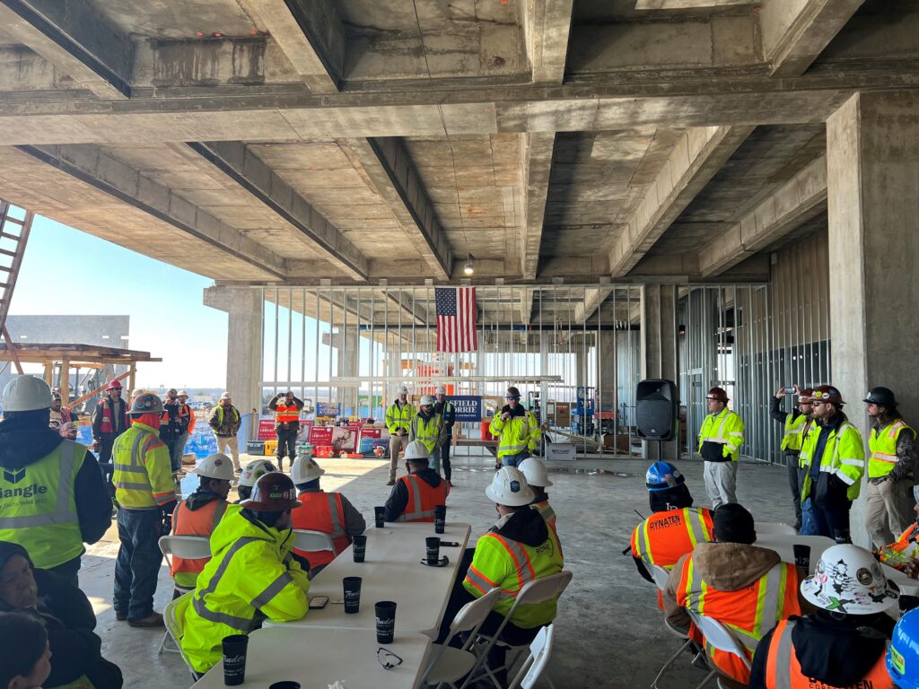 A group of construction workers gather around tables inside of a facility that's under construction. The left side of the facility is open to the outdoors.