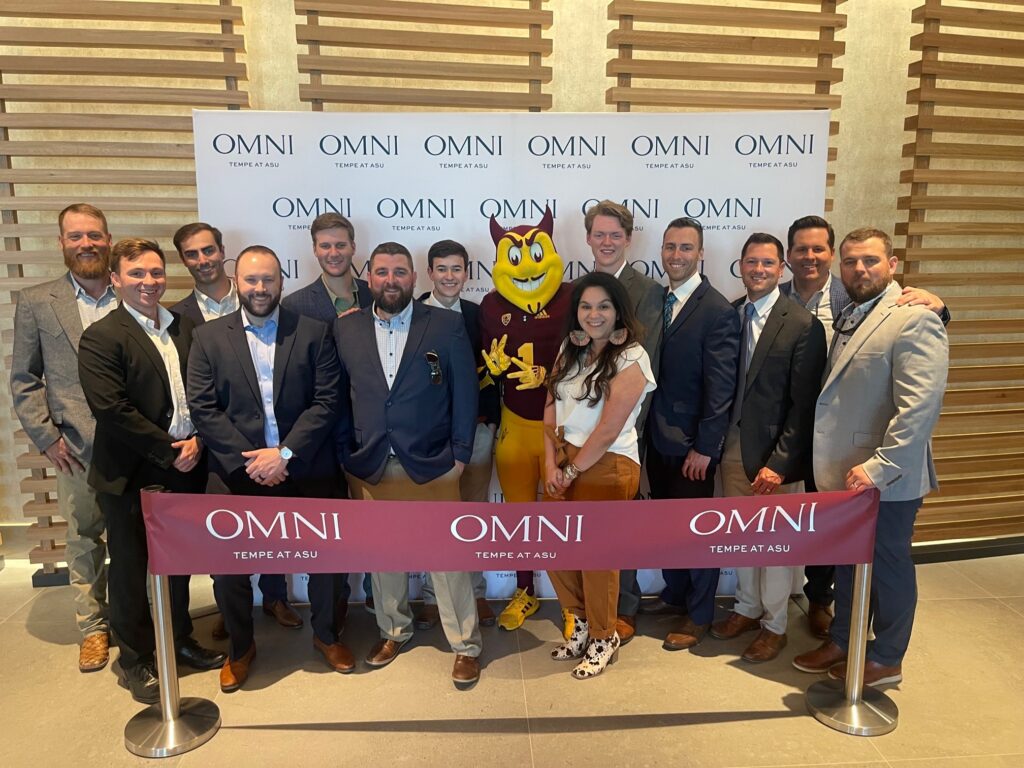 A group of 13 people gather with the Arizona State University mascot, the sun devil. The group is standing in front of an Omni Tempe at ASU backdrop and behind a ribbon that's marked with the same logo.