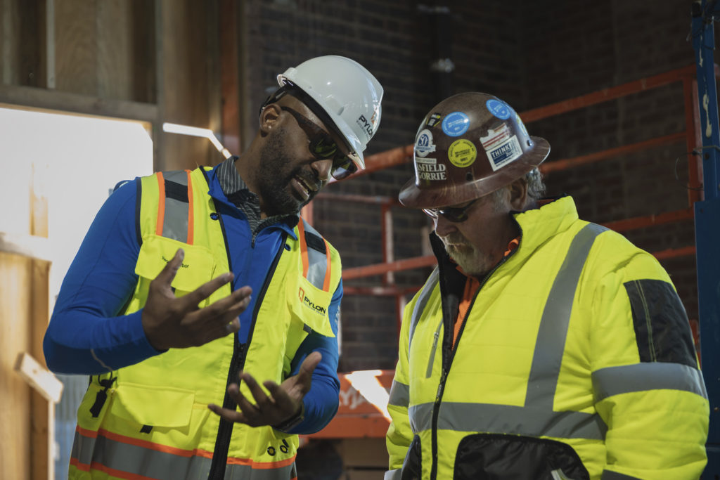 Two men talk while wearing construction personal protection equipment. The man on the left is gesturing as he speaks.