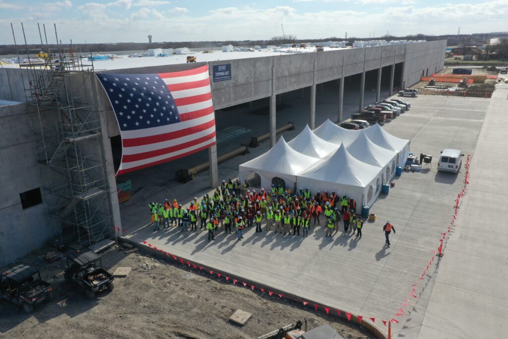 An aerial view of a building under construction. An American flag is attached to the side of the building, and dozens of construction workers in personal protective equipment are gathered in front of white event tents.
