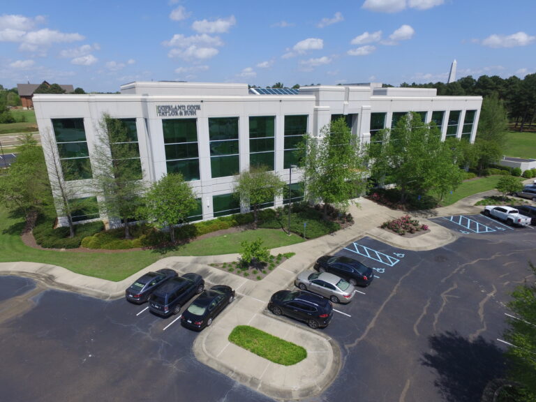Aerial view of a modern multi-story office building with a parking lot and surrounding greenery on a sunny day.