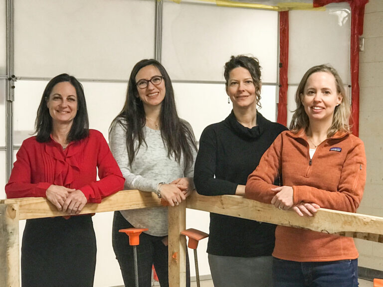 Four women in construction smiling for a photo while standing behind a wooden barrier in a room with exposed pipes in the background.