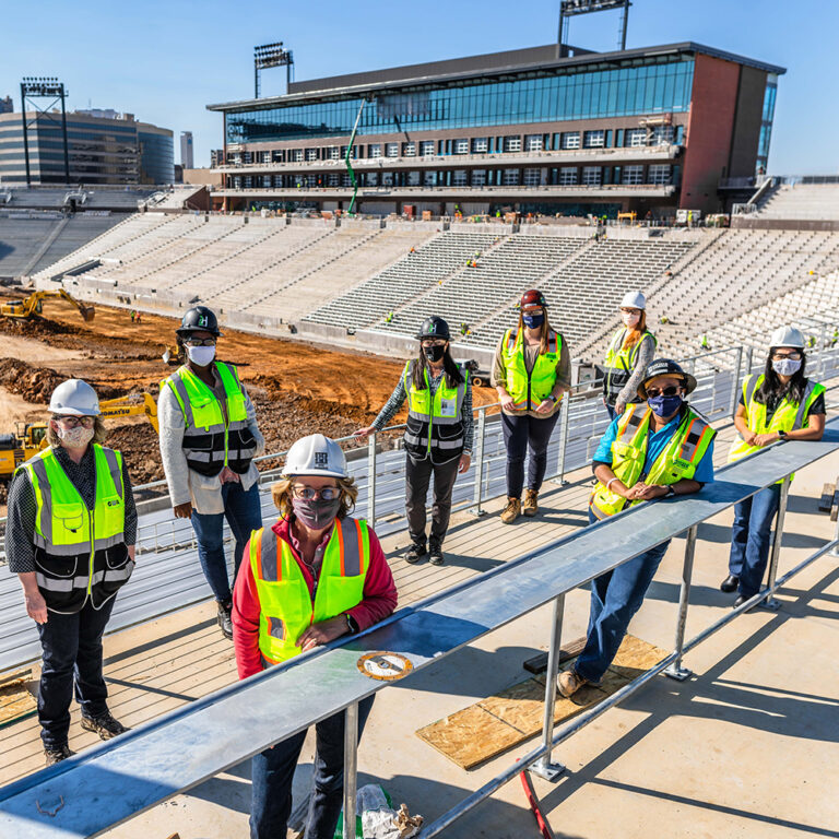 Construction workers in safety gear, including women at work, attending a meeting at a Protective Stadium construction site.