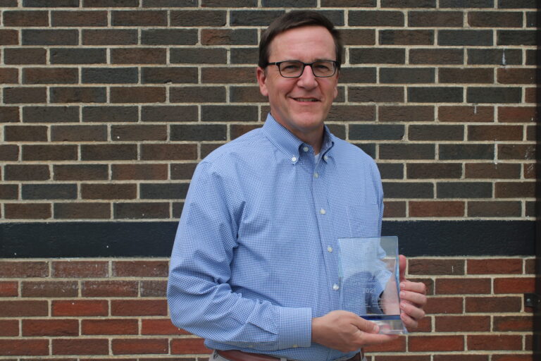 Man in a blue shirt smiling and holding an award in front of a brick wall at Auburn University.
