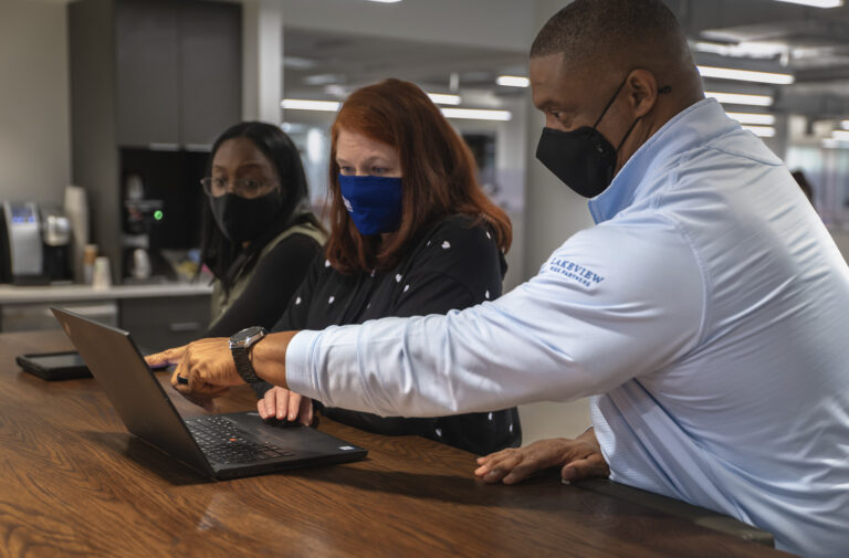 Three individuals in an office setting, two wearing masks, collaborate over a laptop to simplify scheduling.