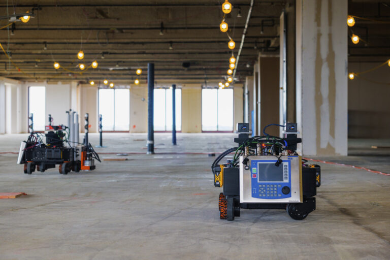 Two robotic devices equipped with various sensors are performing an automated robotic layout in a spacious, unoccupied indoor construction environment.
