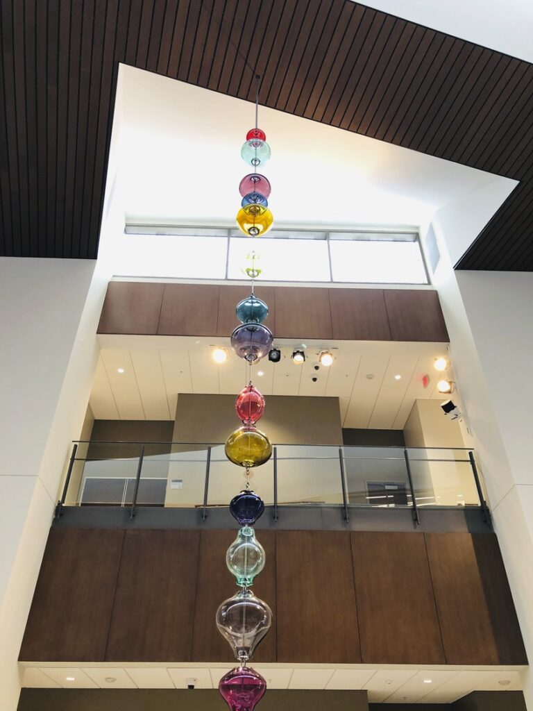 A vertical line of colorful glass orbs suspended in a modern atrium with a wooden ceiling and multiple levels serves as one of the art installations embodying symbols of justice and civic pride.