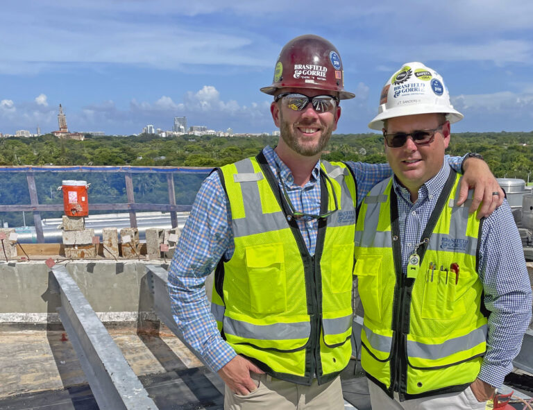 Two construction workers in hard hats and reflective vests posing for a photo with a South Florida construction site in the background.