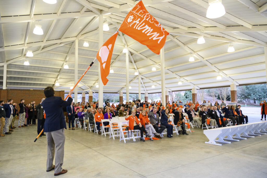 Man speaking to an audience of people dressed in orange at a groundbreaking outdoor event under a shelter.