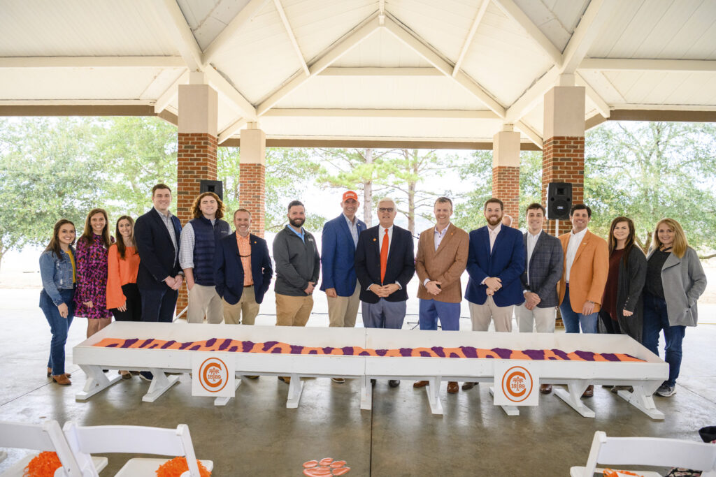 A group of people posing for a photo at an outdoor event with a table in front displaying a branded cloth at the Clemson Alumni and Visitors Center.