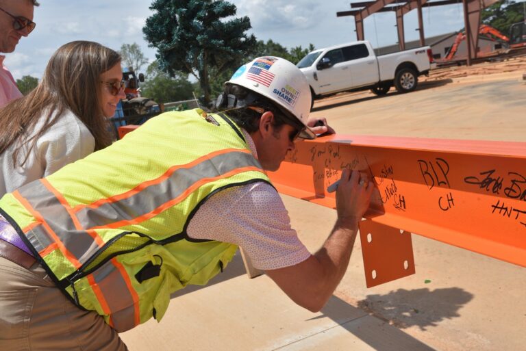 Construction worker in a high-visibility vest signing a steel beam for the Clemson Women's Sports construction milestone with a marker as others look on.