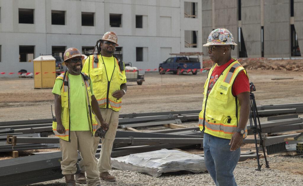 Three construction workers in high-visibility vests and hard hats at a federal project construction site.