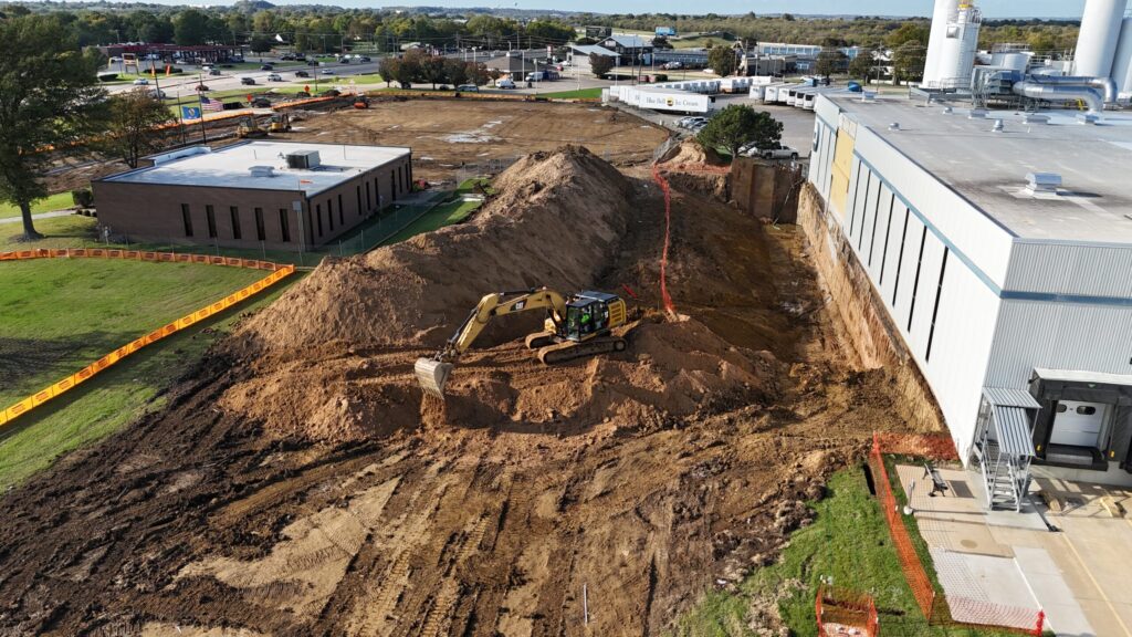 An excavator moving earth for an expansion at a construction site near industrial buildings in Blue Bell, Oklahoma.