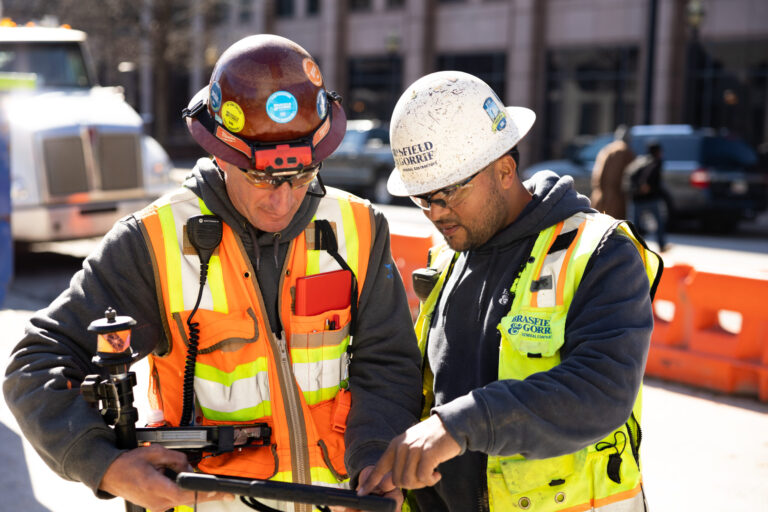 Two construction workers in high visibility vests and hard hats reviewing plans on a clipboard, signifying the start of promising construction careers.
