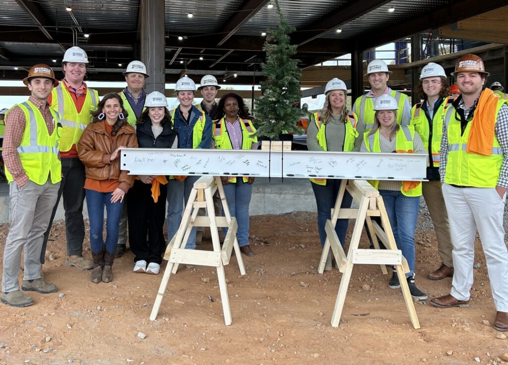 A group of individuals in hard hats and safety vests standing behind a signed steel beam at Clemson University's Alumni & Visitors Center construction site, marking a significant construction milestone.