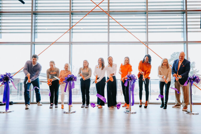 A row of 10 people, mostly women, stand in front of windows to cut a grand opening-type ribbon. The ribbon is purple and the people hold orange scissors--the colors of Clemson University.