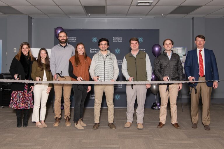 A group of eight people, men and women, stand behind a ceremonial ribbon with scissors in hand, ready for a ribbon cutting.