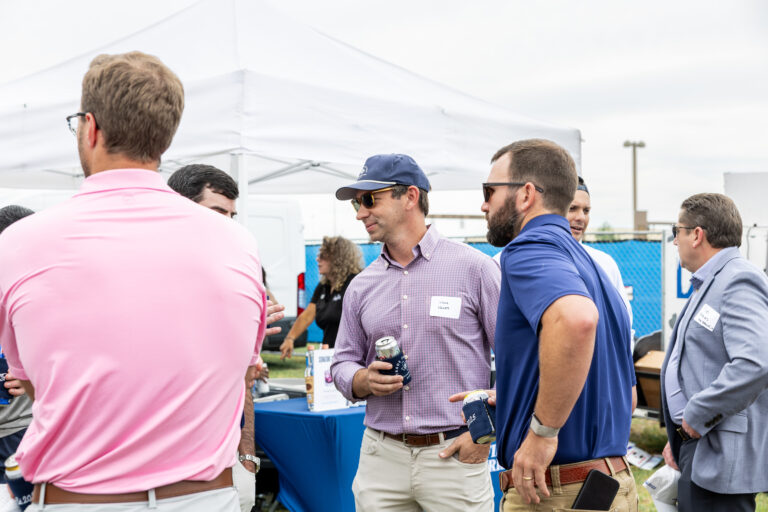 A group of men in casual attire stand and talk near a white canopy tent at an outdoor event, holding drinks and wearing name tags from the Dallas Office of Brasfield and Gorrie.