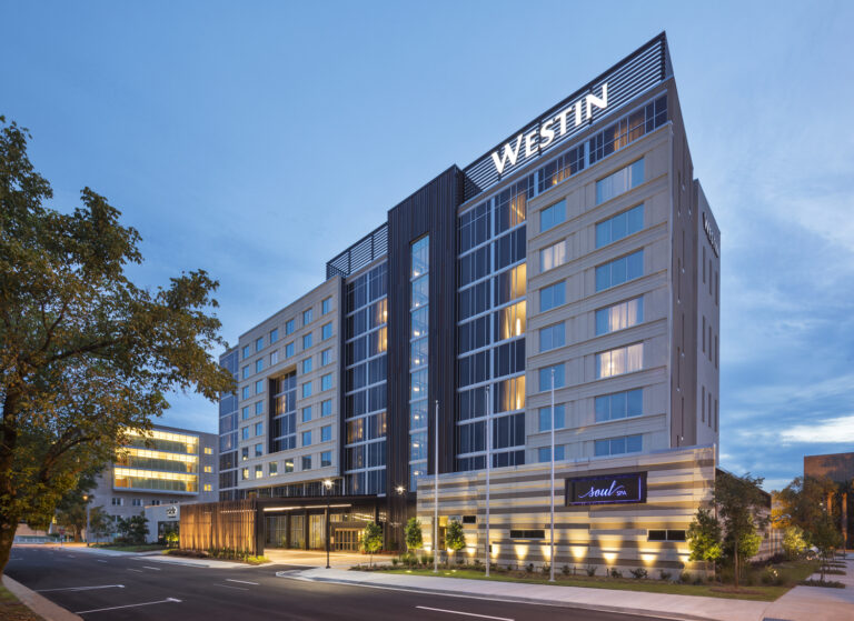 A modern multi-story Westin hotel building by Jackson Office Brasfield and Gorrie, with illuminated signs, large windows, and a tree-lined street at dusk.