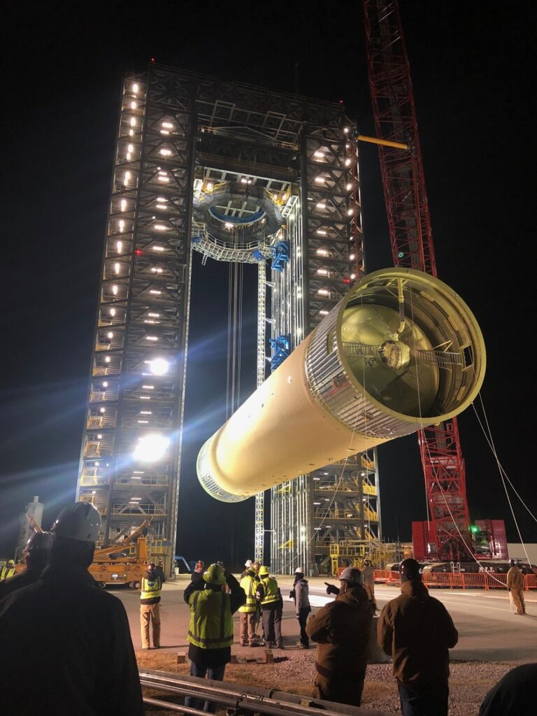 Workers from the Huntsville Office of Brasfield and Gorrie observe as a large cylindrical rocket component is hoisted vertically by a crane within a brightly lit steel structure at night.