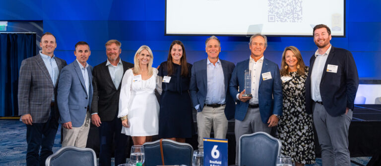 Nine people in business attire stand together, smiling at a formal event in front of a stage with a screen displaying a QR code, representing the Atlanta Office of Brasfield and Gorrie.