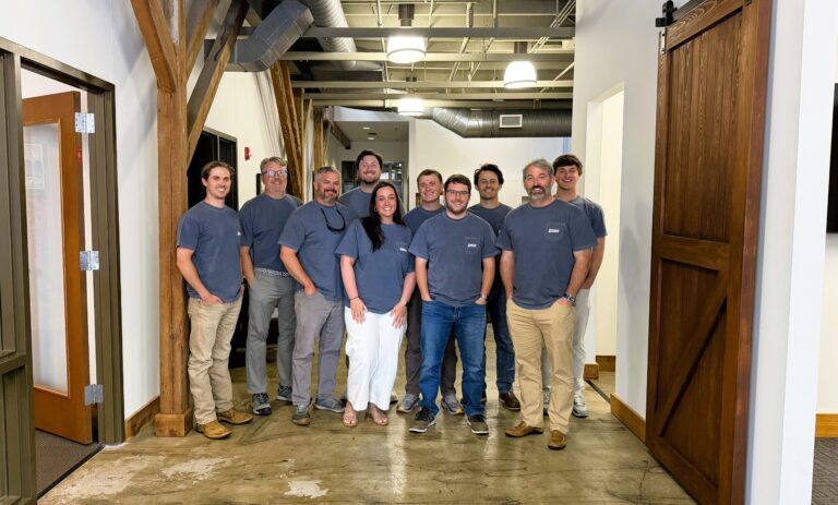 A group of nine people wearing gray shirts and casual pants stand and smile inside the modern Columbus Office of Brasfield and Gorrie, featuring wood accents and exposed ductwork.