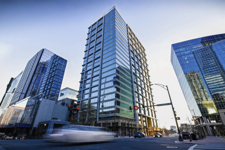 Tall modern office buildings, including the Charlotte Office by Brasfield and Gorrie, rise with sleek glass facades on a bustling city street corner, with cars and traffic lights in the foreground.