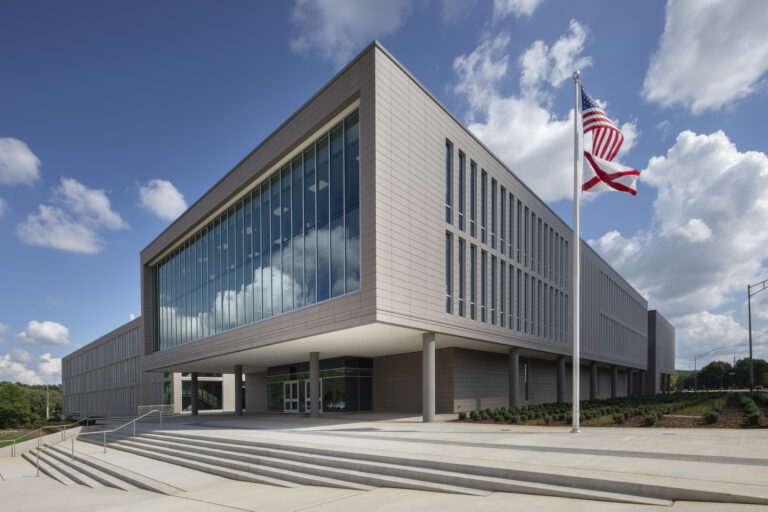 Modern institutional building with large glass windows, two flagpoles displaying the U.S. and Alabama flags, and a wide staircase leading to the Huntsville Office of Brasfield and Gorrie under a partly cloudy sky.