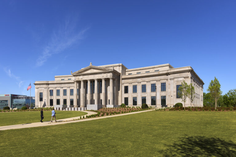 A large neoclassical building with columns at the entrance, surrounded by lawn and landscaping, under a clear blue sky. Two people walk along a path in front of the Huntsville Office Brasfield and Gorrie.
