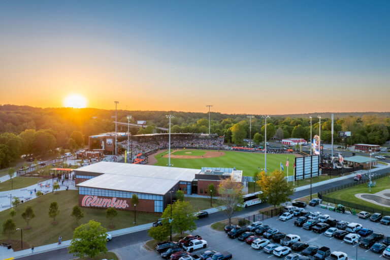 A baseball stadium in Columbus at sunset, with a crowd in the stands, parking lot in the foreground, trees in the background, and the Columbus Office of Brasfield and Gorrie nearby.
