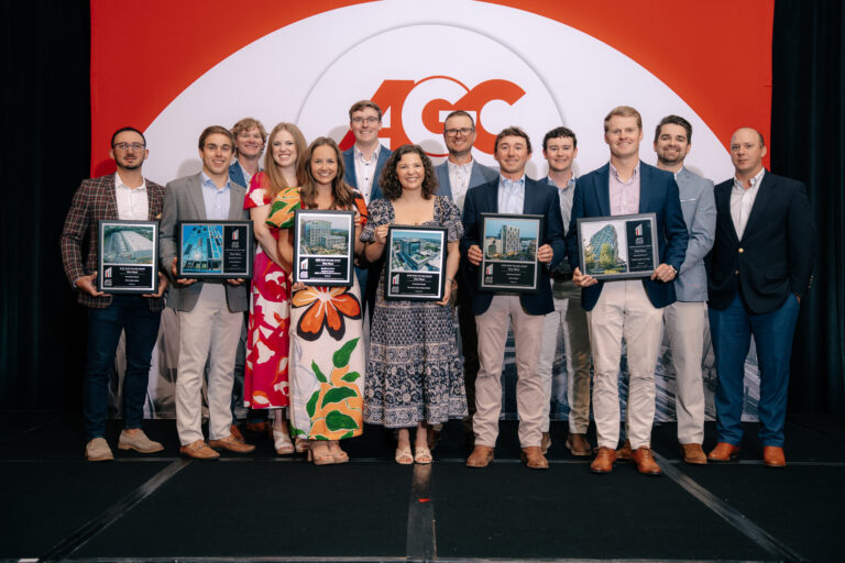 A group of thirteen people from the Atlanta Office of Brasfield and Gorrie stand on stage in business attire, six proudly holding framed awards in front of an AGC logo backdrop.