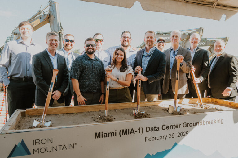Un grupo de personas vestidas con traje de negocios, entre las que se encuentran representantes de la oficina de Miami de Brasfield and Gorrie, posan con palas en el acto de inauguración de las obras del centro de datos de Iron Mountain en Miami, celebrado el 26 de febrero de 2025. Al fondo se ve una excavadora.