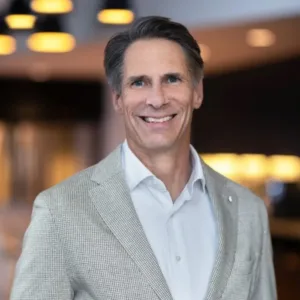 A man in a light gray blazer and white shirt smiling in a modern indoor setting with blurred lights in the background.