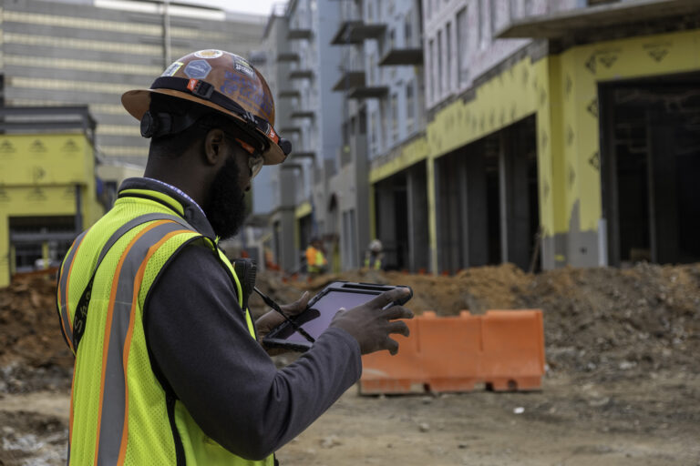A construction worker holding a digital tablet in front of a construction site.