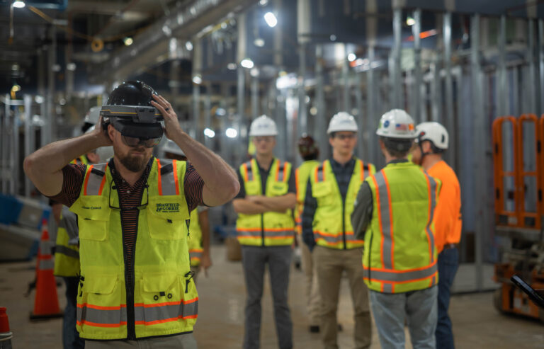 A group of construction workers wearing safety vests while the worker in the foreground adjusts his helmet.