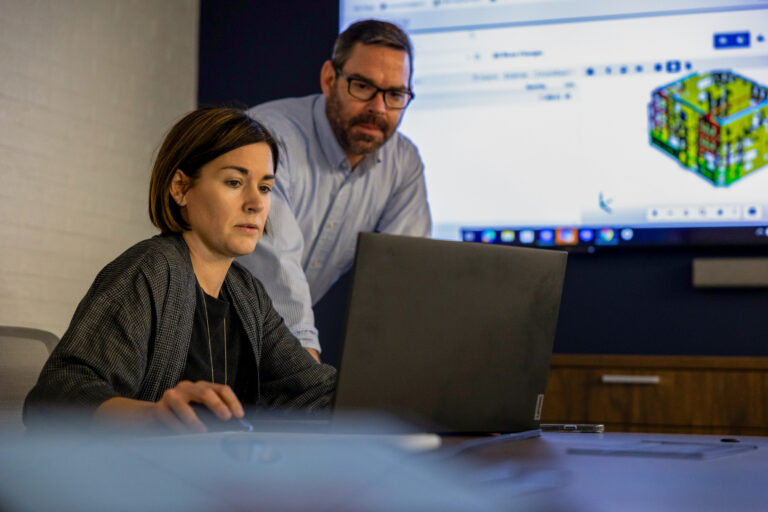 Two professionals analyzing data on a laptop in an office setting with a data visualization projected on the screen in the background.