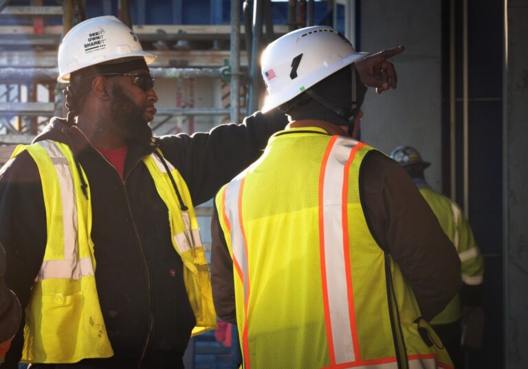 Three construction workers wearing hard hats and safety vests, one in hte foreground points out of frame