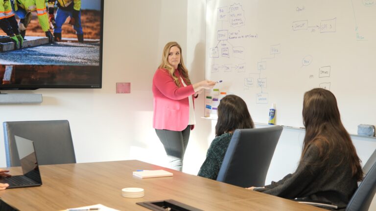 A woman presenting in a meeting room with attentive colleagues.