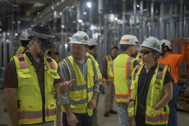 Group of industrial workers wearing safety gear having a discussion in a factory setting, one wears a set of AR goggles