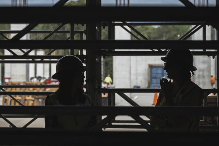 Two silhouetted construction workers in hard hats and reflective vests having a conversation at a construction site.