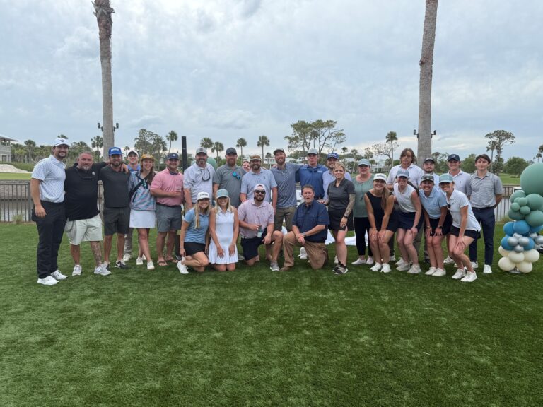 Un grupo de la oficina de Jacksonville de Brasfield and Gorrie posa al aire libre sobre un césped, vestido con ropa informal y de golf, con palmeras y un cielo nublado al fondo.