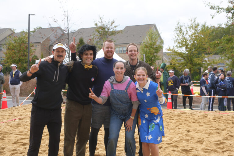 Six adults from the Atlanta Office of Brasfield and Gorrie pose and smile together on a sand court outside, some wearing costumes, with houses and spectators visible in the background.