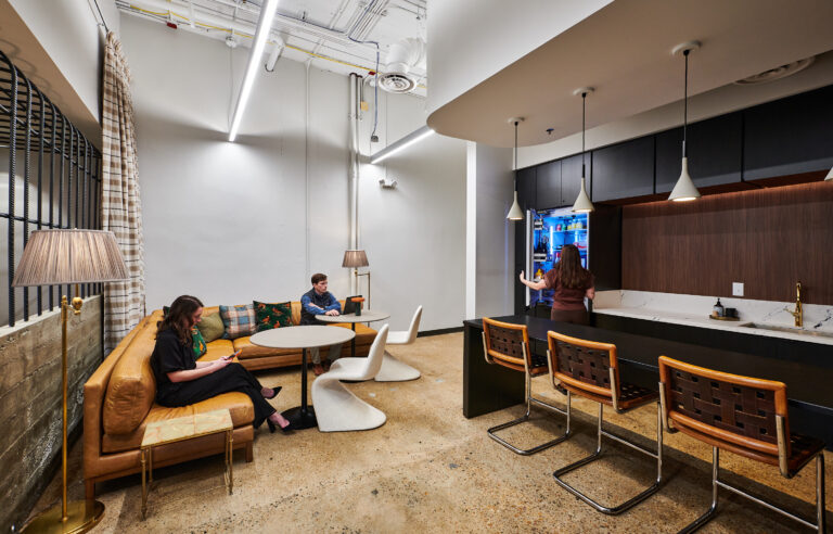 Three people are in the modern Huntsville office lounge of Brasfield and Gorrie; two are seated at a table and on a sofa, while one stands by a kitchen area with bar stools and a refrigerator.