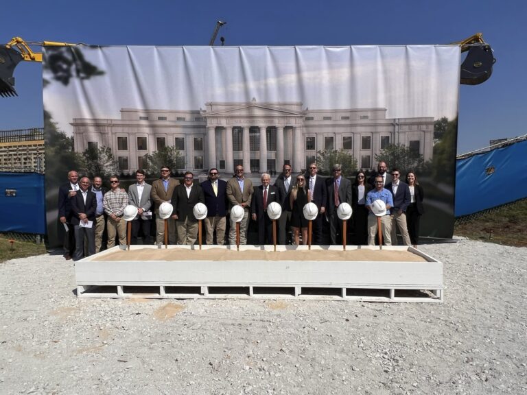 Group of individuals in business attire and hard hats standing in front of a construction site banner.